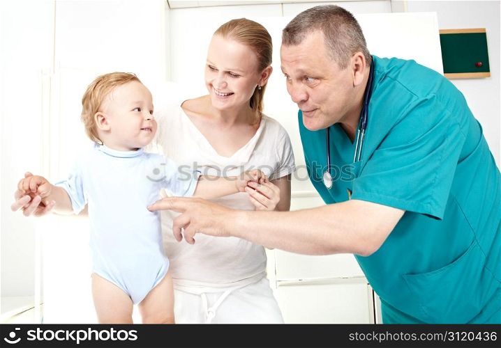 A doctor of 45-50 years old in a green smock is playing with a 1,5 year-old boy in a light medical study. The young mother of 25-30 in white clothes is looking at her son with a smile.