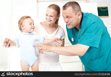 A doctor of 45-50 years old in a green smock is playing with a 1,5 year-old boy in a light medical study. The young mother of 25-30 in white clothes is looking at her son with a smile.