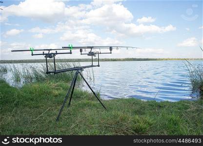 A double fishing rod stand with cast rods in place stand in the green grass alongside the calm waters of the dam,
