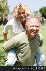 A elderly gentleman gives a piggyback to a lady outdoors in a park on a summers day