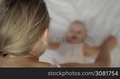 A fair-haired woman doing some playful exercises with her six months old baby daughter who is lying in front of her on white bed linen. Woman is holding baby&acute;s arms with her hands and moving them from side to side. Even though baby is a bit blurred, it is obvious that she is having good time