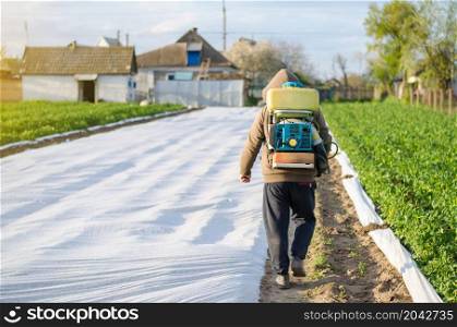 A farmer with a mist sprayer on his back walks through the farm field. The use of chemicals for crop protection in agriculture. Protection of cultivated plants from insects and fungal infections.