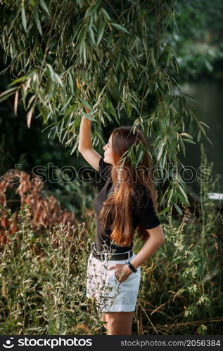 A fascinating girl among willow leaves on the shore of a pond.. A stylish beauty in a black T-shirt on the shore of a pond 3653.