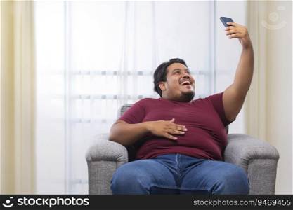  A fat man happily clicking a selfie in his mobile while sitting on couch.
