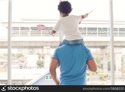 A father carrying his african black little son while a boy waving american flag beside the window. Diversity and black lives matter concept.