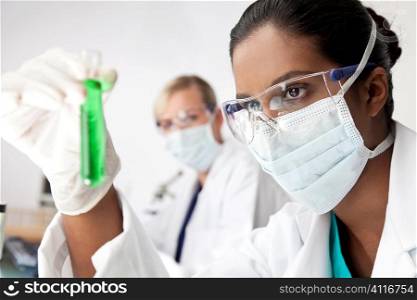 A female Asian medical or scientific researcher or doctor looking at a test tube of a green solution in a laboratory with her blond female colleague out of focus behind her.