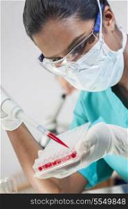 A female Asian medical or scientific researcher or doctor using a pipette and sample tray to test blood sample in a laboratory with her colleague out of focus behind her.