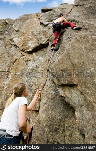 A female belaying a male on a steep rock face. Shallow depth of field with the focus on the climbing which is belaying (the on at the bottom)