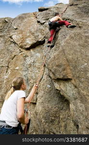 A female belaying a male on a steep rock face. Shallow depth of field with the focus on the climbing which is belaying (the on at the bottom)