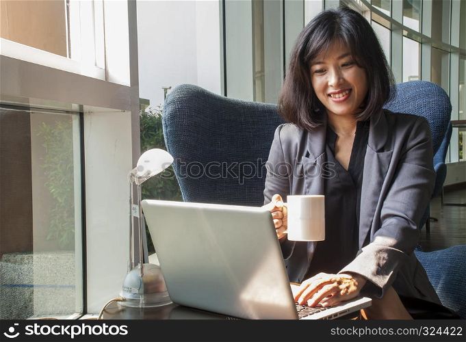A female businessman is smiling while working with a computer and using the right hand to hold a coffee for drinking in her office.