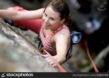 A female climber on a steep rock face viewed from above with the belayer in the background. The climber is smiling at the camera. Shallow depth of field is used to isolated the climber.