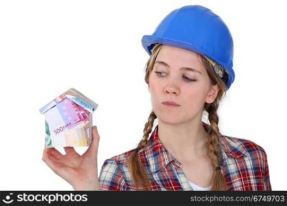 A female construction worker holding a miniature of a house made of bills.
