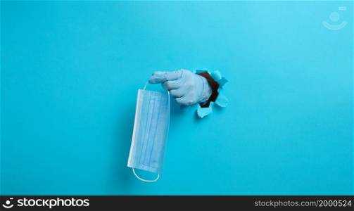 a female hand holds a disposable medical mask to protect against viruses during an epidemic and pandemic. Part of the body sticks out of a torn hole in blue paper