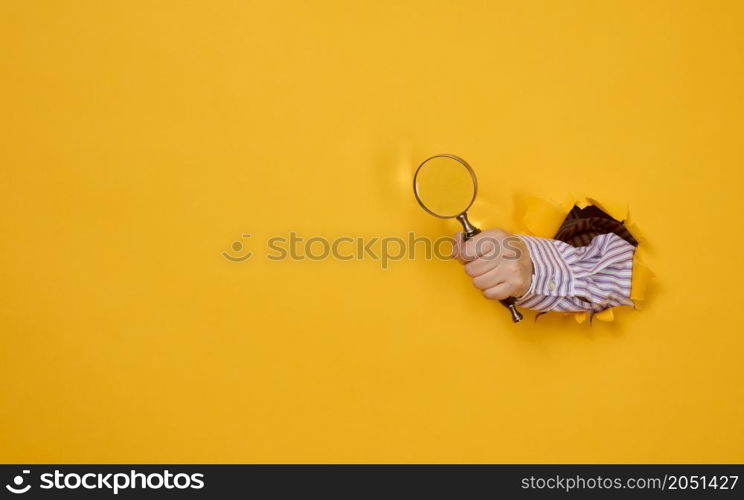 a female hand holds a magnifying glass on a yellow background, part of the body sticks out of a torn hole in a paper background. Business concept, finding information, answering questions