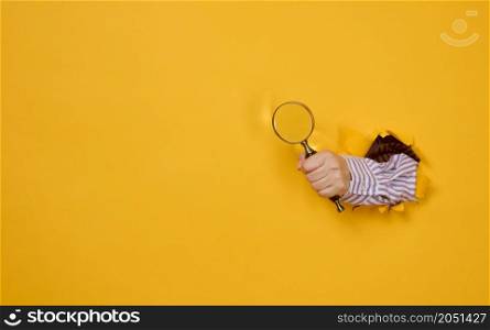 a female hand holds a magnifying glass on a yellow background, part of the body sticks out of a torn hole in a paper background. Business concept, finding information, answering questions
