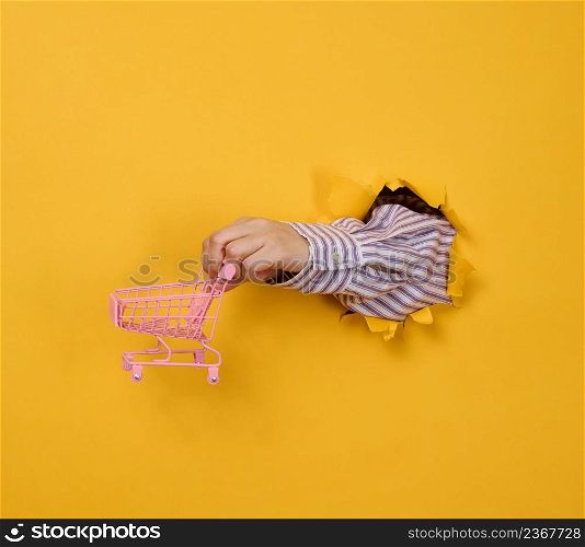 a female hand holds a miniature shopping cart on a yellow background, a part of the body sticks out of a torn hole in a paper background. Business concept, start of sales and online shopping