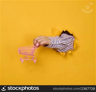 a female hand holds a miniature shopping cart on a yellow background, a part of the body sticks out of a torn hole in a paper background. Business concept, start of sales and online shopping