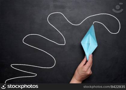 A female hand holds a paper boat against a black chalkboard background with a long path. Concept of mentorship and assistance