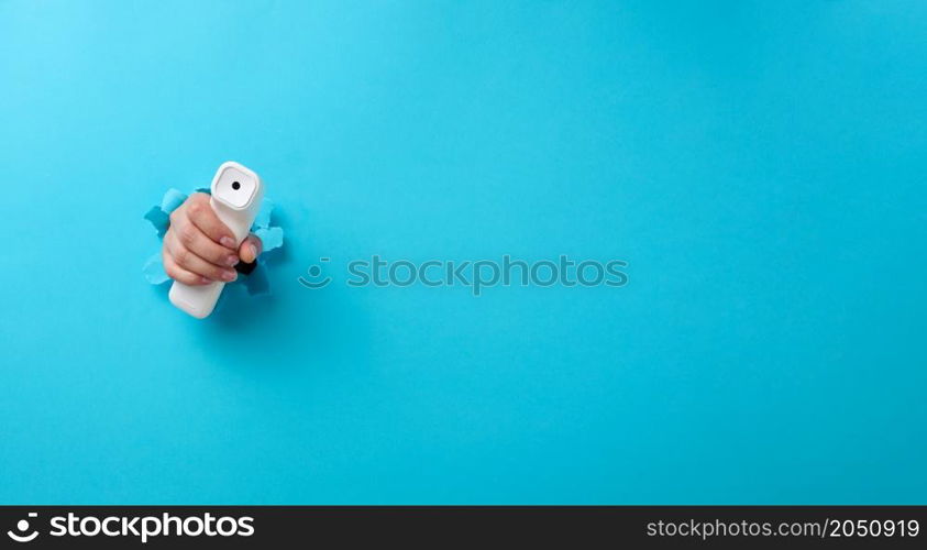 a female hand holds a plastic non-contact thermometer to measure a person&rsquo;s temperature, a part of the body sticks out of a torn hole in a blue paper background. Copy space