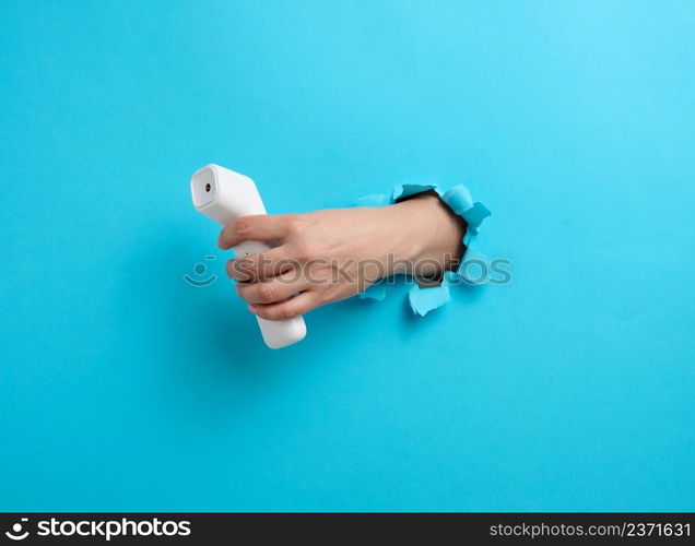 a female hand holds a plastic non-contact thermometer to measure a person&rsquo;s temperature, a part of the body sticks out of a torn hole in a blue paper background