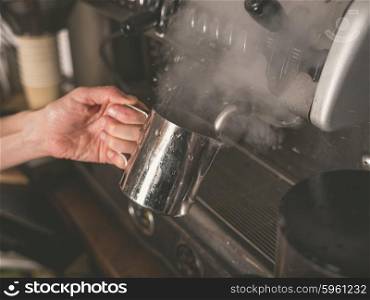 A female hand is placing a cup under the froth element of a professional coffee machine which causes steam to rise from the liquid in the cup