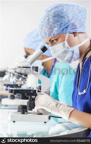A female medical or scientific researcher or doctor using her microscope in a laboratory with her Asian colleague out of focus behind her.