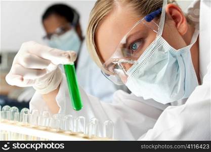A female medical or scientific researcher or doctor using looking at a green solution in a laboratory with her Asian female colleague out of focus behind her.