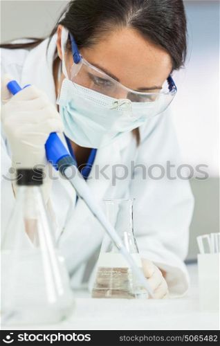 A female medical or scientific researcher or scientist using a pipette and flask in a laboratory with a computer monitor behind her