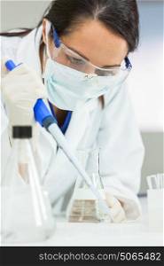 A female medical or scientific researcher or scientist using a pipette and flask in a laboratory with a computer monitor behind her