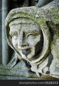 A figure of a weathered gargoyle on the belltower of the Church of St. Mary The Virgin in Oxford, England. The carving appears to be a monk in a robe and cowl.
