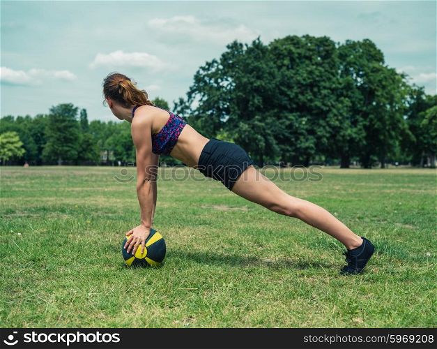 A fit and athletic young woman is doing a plank exercise on a medicine ball in the park on a sunny summer day