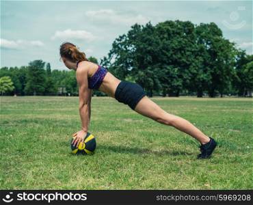 A fit and athletic young woman is doing a plank exercise on a medicine ball in the park on a sunny summer day