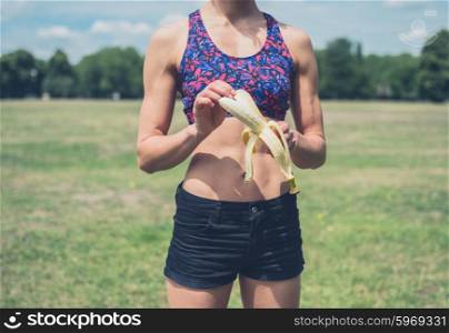 A fit and athletic young woman is peeling a banana in the park on a sunny summer day