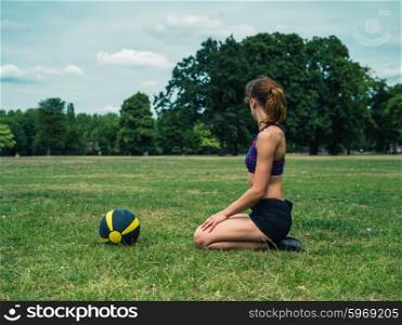 A fit and athletic young woman is sitting on the grass in a park with a medicine ball in front of her