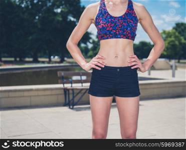 A fit and athletic young woman is standing in a park by some benches on a sunny summer day
