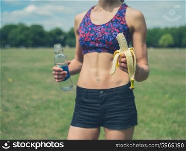 A fit and athletic young woman is standing in the park with a banana and a water bottle
