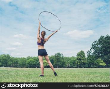 A fit and athletic young woman is standing on the grass in a park with a hula hoop raised above her head