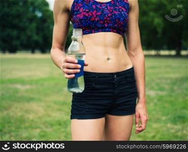 A fit and athletic young woman is standing on the grass in a park with a water bottle in her hand