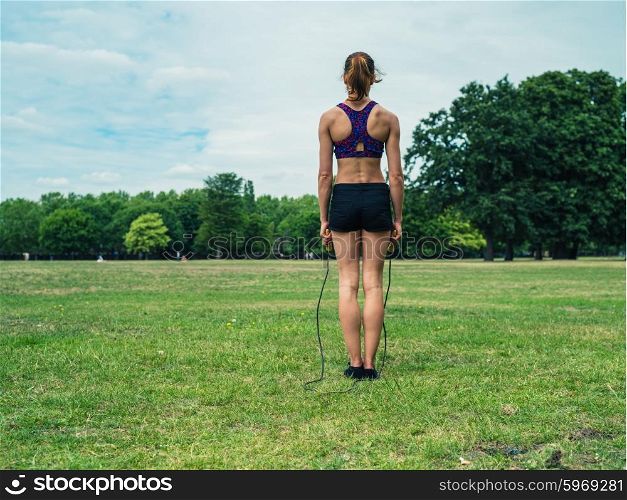 A fit and athletic young woman is standing on the grass in a park and is exercising with a jump rope