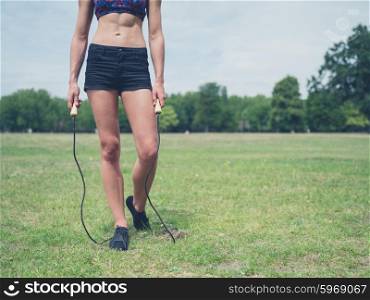 A fit and attractive young woman is standing on the grass in a park with a jump rope on a sunny day of summer