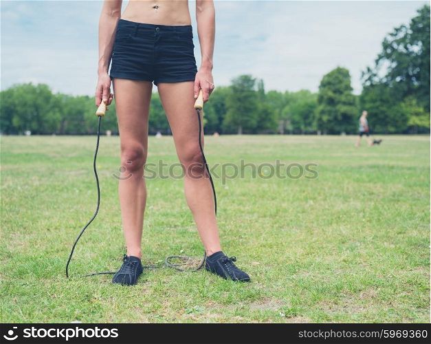 A fit and attractive young woman is standing on the grass in a park with a jump rope on a sunny day of summer