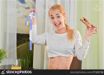 A fit, blonde woman smiles happily while holding a measuring tape and a sandwich on an out of focus background. Healthy lifestyle concept.. Blonde woman is happy about weight loss