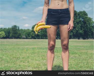 A fit young woman is standing on the grass in a park with two bananas in her hand