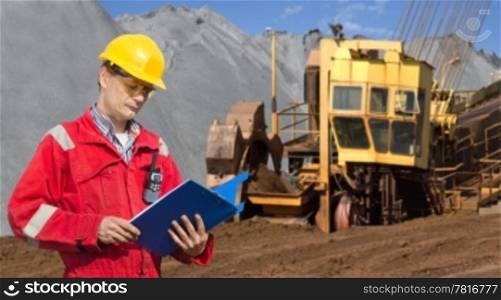 A foreman in a mining site, checking the logs in a blue folder, with a huge wheel digger in the background