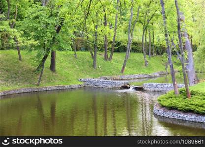 A forest lake with a cascade overflow of water in a beautiful park is located among various deciduous trees, framed on the banks of connected cobblestones, green grass and clipped shrubs under soft sunlight.. A forest lake surrounded by trees in a beautiful park, framed by cobblestone shores in the rays of soft light.
