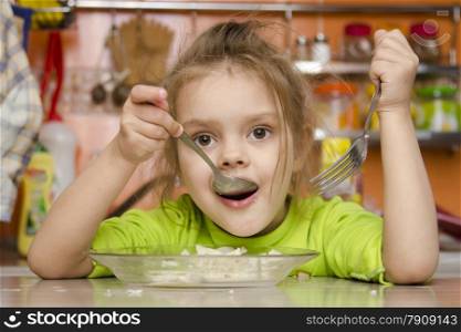A four year old girl eats sitting at the table in the kitchen. In one arm and the girls spoon in the other fork. Girl eat with a spoon
