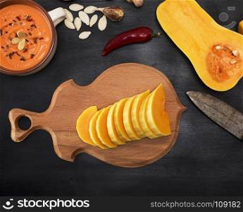 A fresh pumpkin is sliced into pieces on a kitchen cutting board and a plate with pumpkin soup, top view