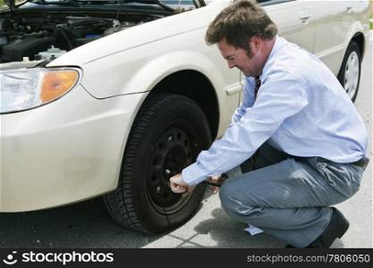 A frustrated businessman loosening lug nuts on his car&rsquo;s flat tire.