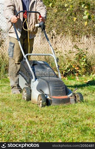 A gardener mows grass with an electric lawn mower in his backyard on a bright sunny fall day. Vertical image. Copy space.. A gardener mows green grass with an electric lawn mower on a bright sunny autumn day.