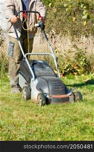 A gardener mows grass with an electric lawn mower in his backyard on a bright sunny fall day. Vertical image. Copy space.. A gardener mows green grass with an electric lawn mower on a bright sunny autumn day.